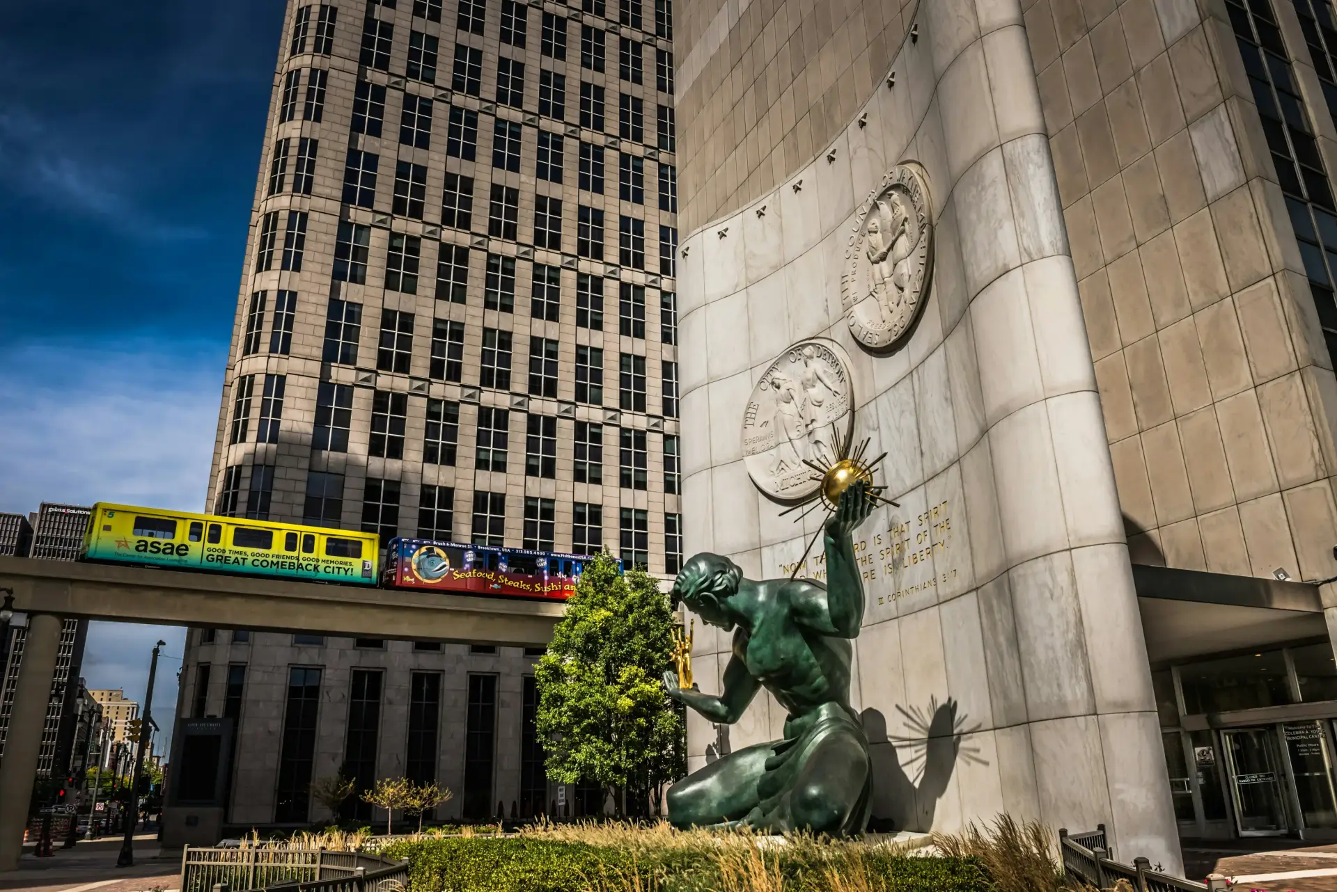 The Spirit of Detroit bronze statue sits in front of a marble wall at the Coleman A. Young Municipal Center, with a yellow Detroit People Mover train crossing an elevated track in front of a tall skyscraper in the background.